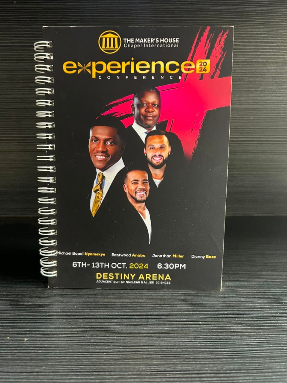 Front of a black wire-bound conference notebook showing four men in suits, a gold conference title with the year, the dates 6 to 13 October 2024 and the venue name.