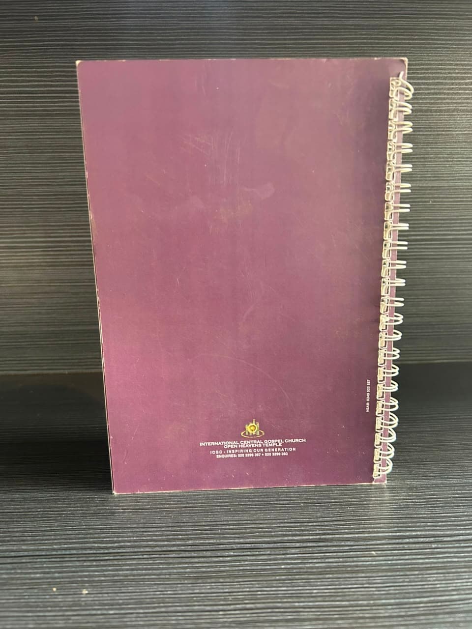 The back of a plum wire-bound notebook with a small gold crest and a line of contact details near the bottom, standing on a dark shelf.